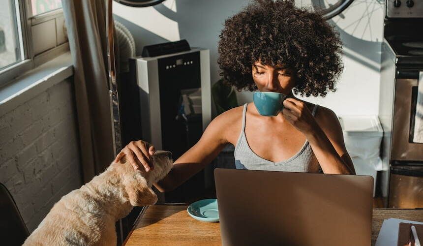 Junge Frau mit dunklen Locken sitzt an einem Tisch mit Laptop, trinkt aus einer türkisfarbenen Tasse und streichelt einen Hund dabei